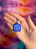 Hand holding a blue keychain with a square logo against a tie-dye background