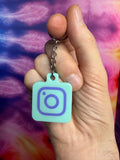 Hand holding a light blue keychain with a purple Instagram logo against a colorful tie-dye background