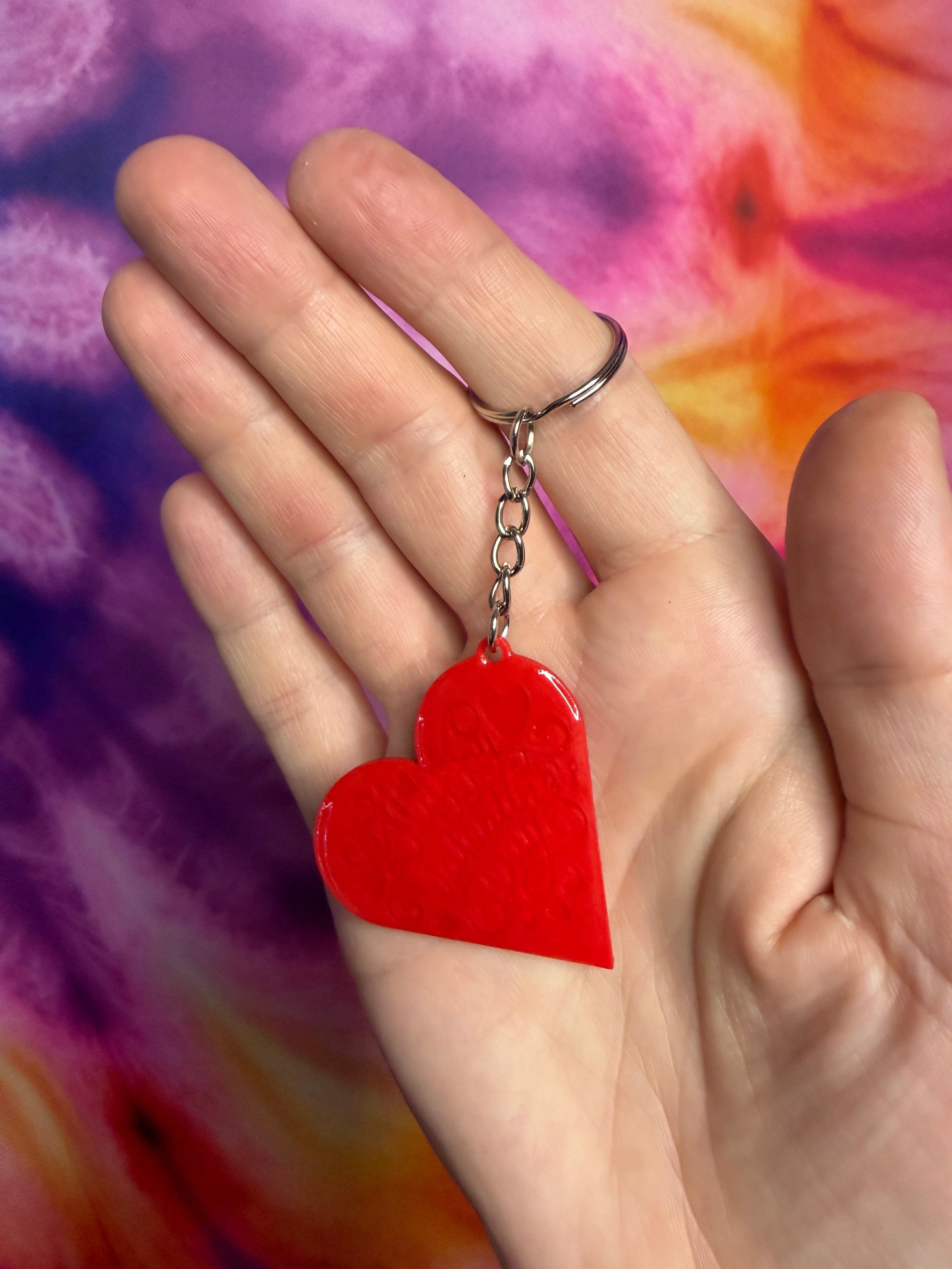 Red heart-shaped keychain Happy Valentines Day held in a hand against a colorful tie-dye background
