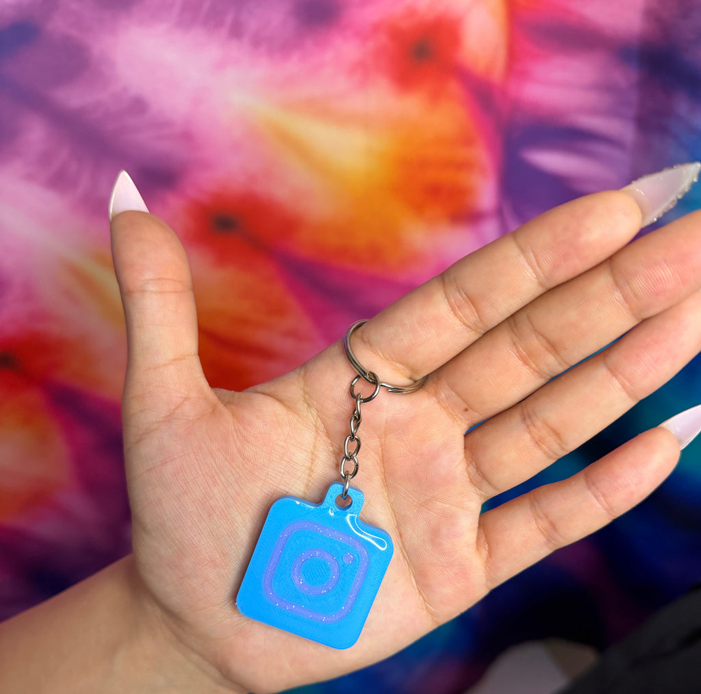 Hand holding a blue keychain with a camera design against a tie-dye background