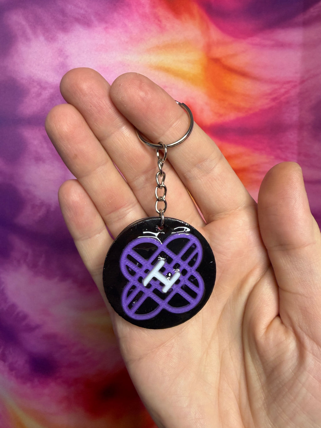 Keychain with a purple k pop demon hunter huntrix symbol held in a hand against a tie-dye background