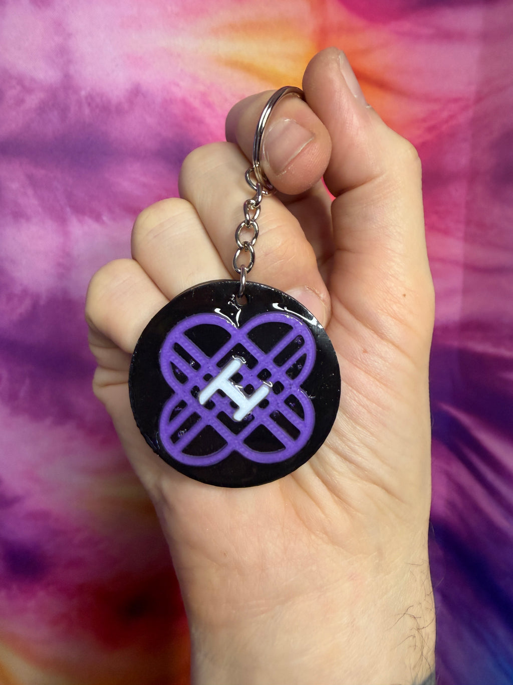 Hand holding a keychain with a purple k pop demon hunter huntrix  symbol against a tie-dye background