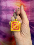 Yellow gold and pink keychain with Instagram logo held in a hand against a tie-dye background