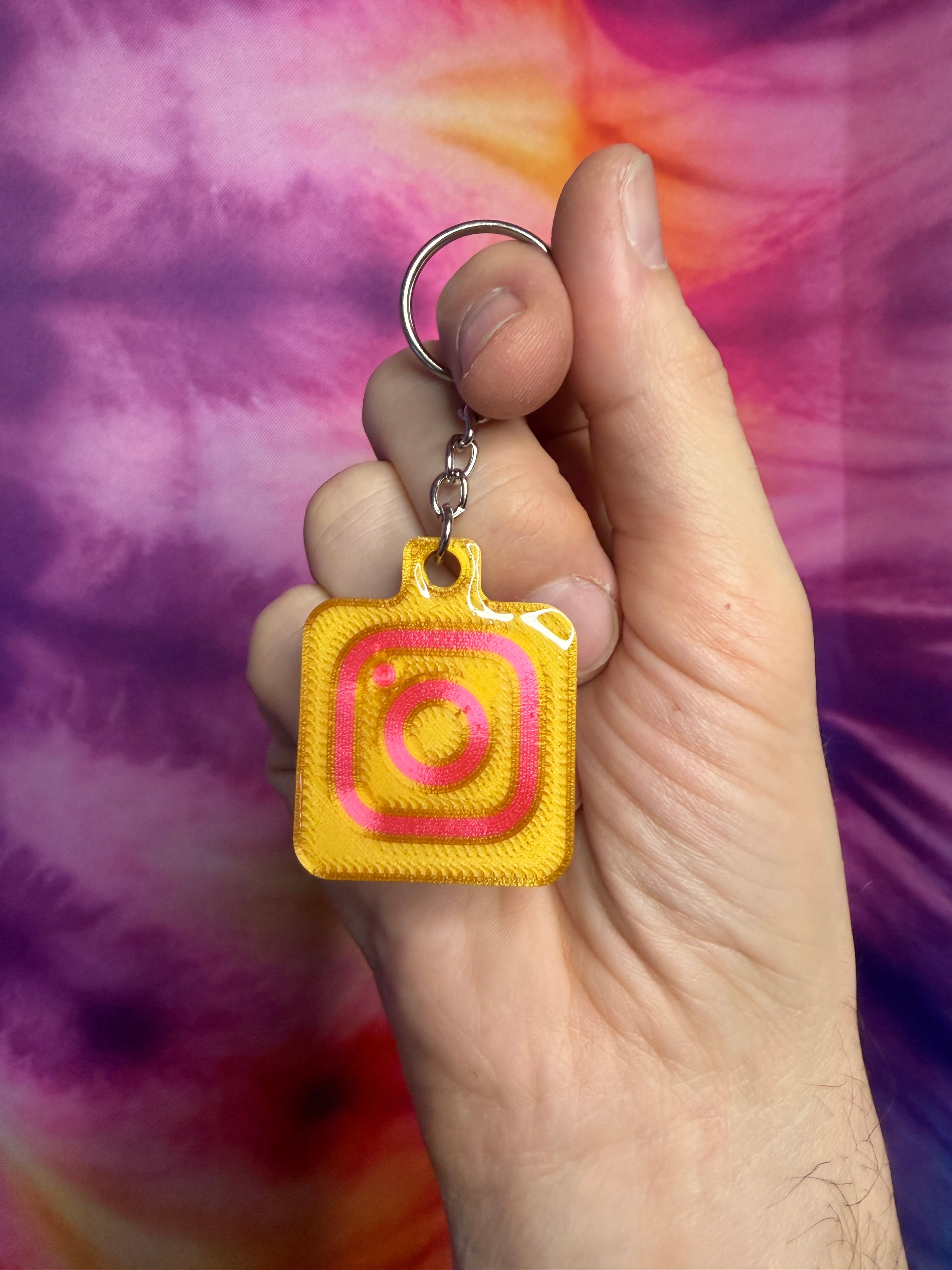 Yellow gold and pink keychain with Instagram logo held in a hand against a tie-dye background
