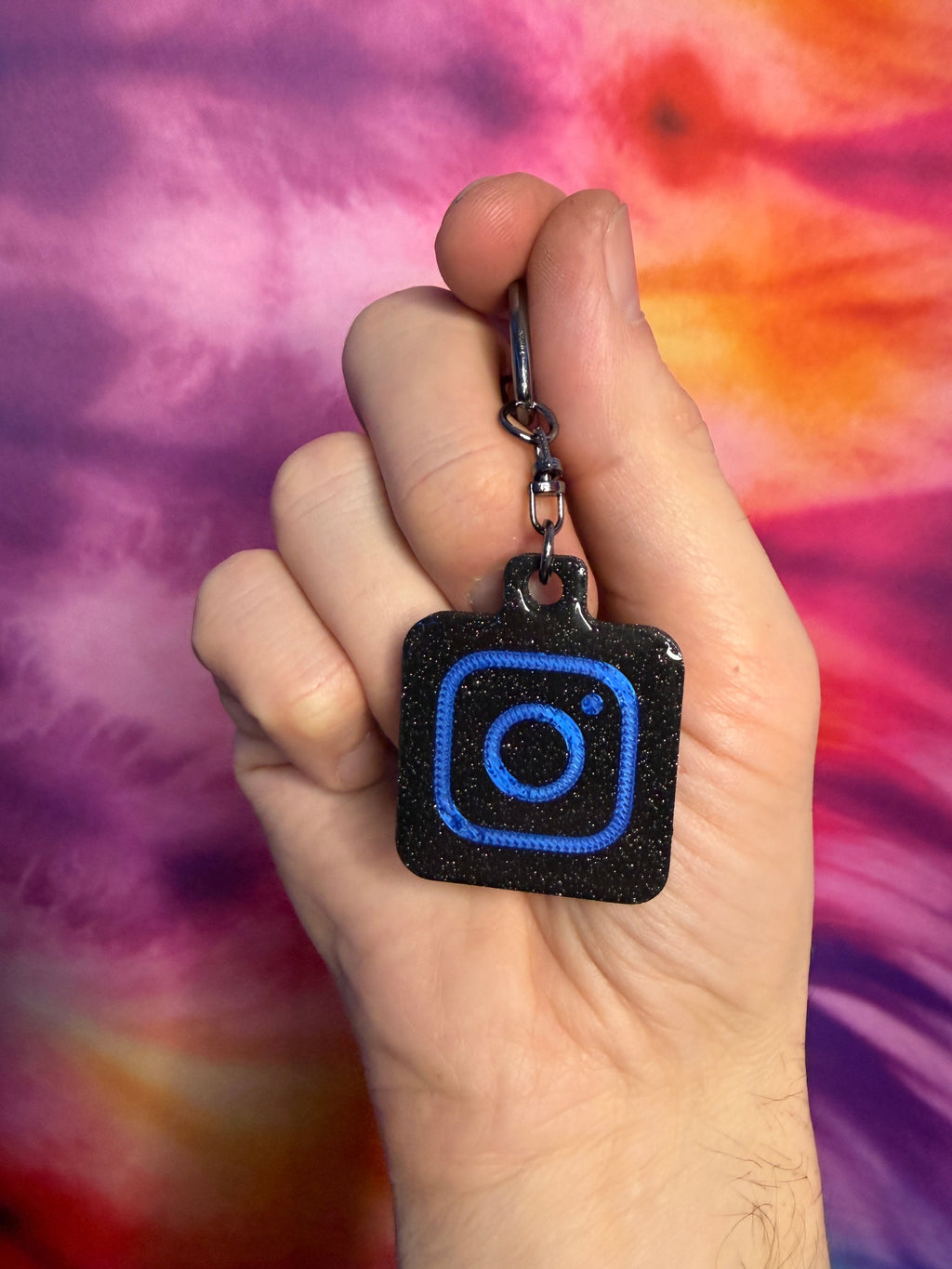 Hand holding a keychain with a blue Instagram logo against a tie-dye background
