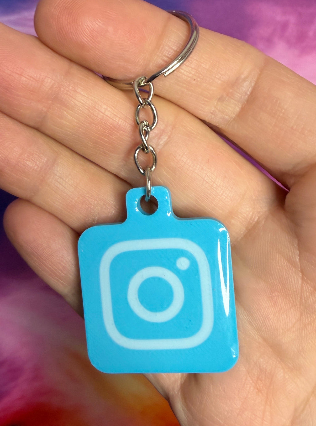 Hand holding a blue Instagram logo keychain against a tie-dye background
